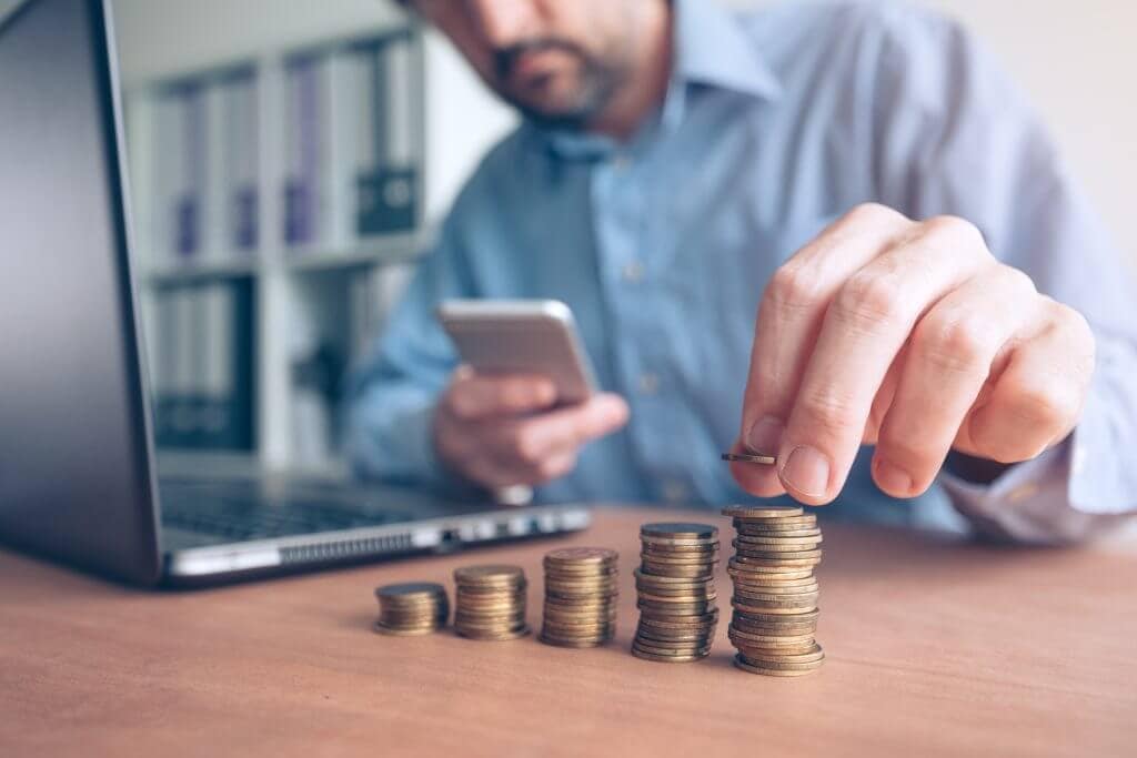 a man organizing a few stacks of coins next to his laptop after checking his phone